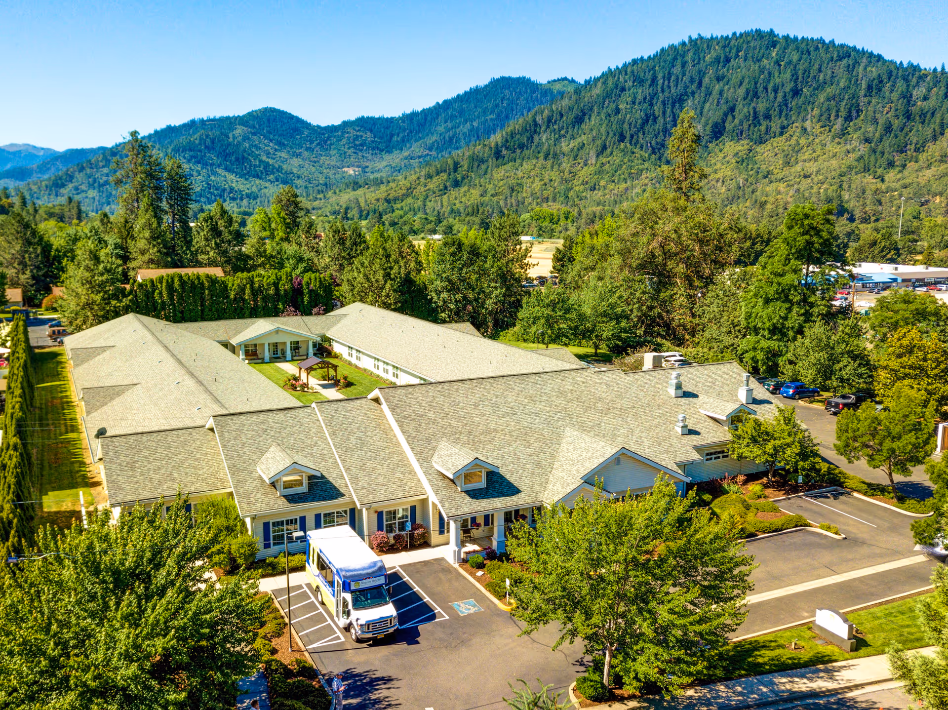 Aerial view of Morrow Heights Assisted Living facility surrounded by trees and greenery with mountains in the background. The building has a light-colored roof and a courtyard in the center. A white shuttle bus is parked in front of the entrance, and there are several parking spaces and cars visible around the building.