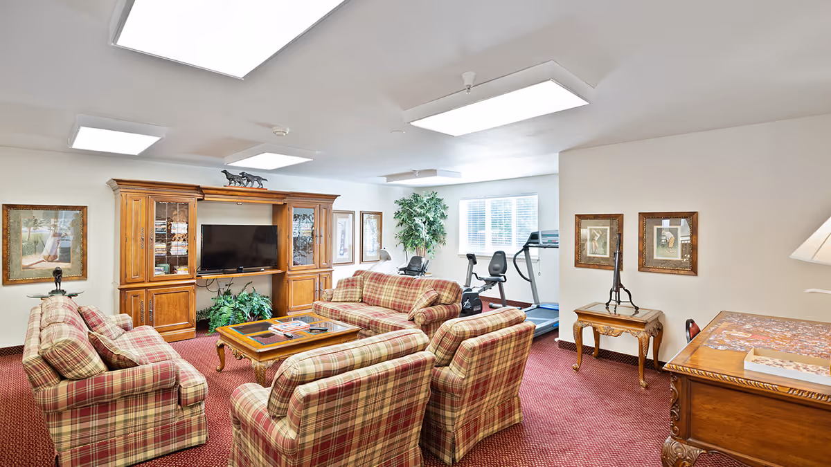 A cozy living room area with plaid upholstered sofas and armchairs arranged around a wooden coffee table. A large wooden entertainment center with a flat-screen TV and decorative items is against the wall. The room has framed artwork, a potted plant near a window, and exercise equipment including a treadmill and stationary bike in the background. The carpet is red, and the walls are light-colored with ceiling lights providing bright illumination.