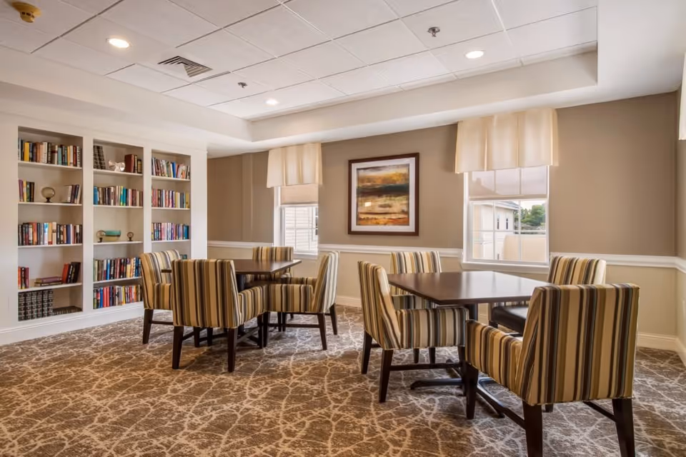 A cozy interior room with two square wooden tables surrounded by striped upholstered chairs. The room features a built-in bookshelf filled with books, two windows with cream-colored valances, a framed abstract painting on the wall, and patterned carpet flooring.