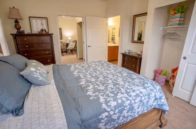 Bedroom with a bed dressed in blue floral bedding, wooden dressers, an open closet, and a doorway leading to a bathroom and sitting area.