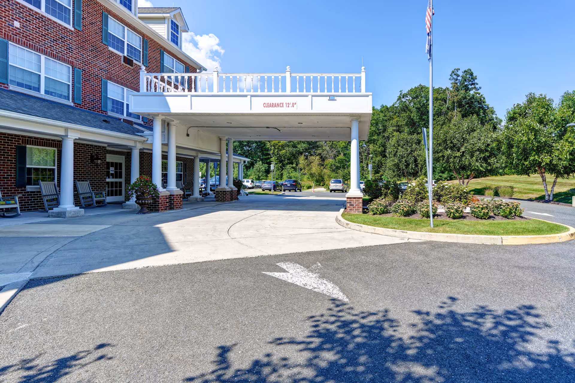 Covered entrance and porte-cochère of a brick senior living building with rocking chairs, circular driveway, flagpole, and landscaped grounds.