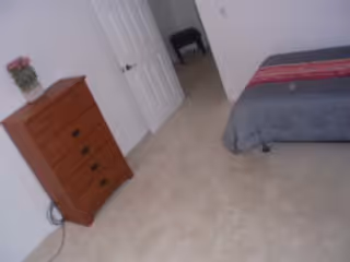 Carpeted bedroom with a partially visible bed on the right, a wooden dresser with flowers on the left, and an open door leading to a hallway.
