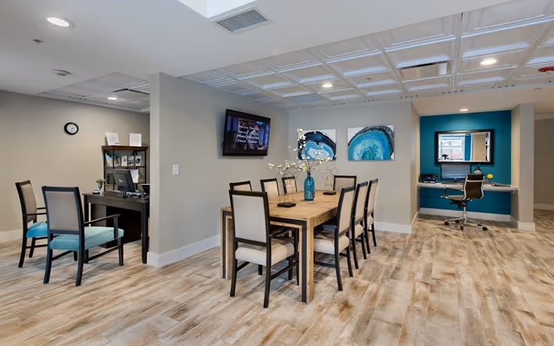 Bright communal interior showing a long wooden dining table with chairs, a wall-mounted TV, artwork, and adjacent workstations.