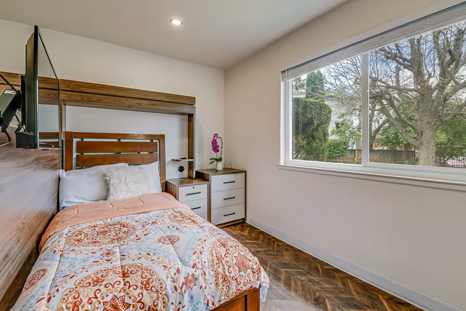 Single-occupancy bedroom with a patterned comforter, wooden headboard, nightstand and a large window overlooking trees.