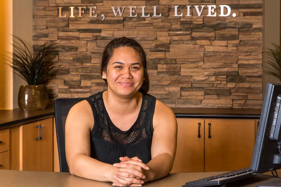 A woman with dark hair tied back is sitting at a reception desk with her hands clasped, smiling. Behind her is a stone wall with the words 'LIFE, WELL LIVED.' displayed. There are cabinets and a plant on the left side, and a computer monitor on the right side of the desk.
