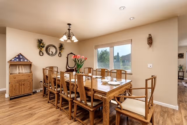 A dining room with a long wooden table set for eight people with plates and bowls. The room has wooden chairs with cushions, a wooden floor, and beige walls. There is a window with a view of greenery outside, a chandelier hanging from the ceiling, and decorative items on the walls including a round mirror and floral arrangements. A wooden cabinet with a folded American flag in a triangular display case is visible in the corner.