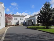 Exterior view of a senior living facility building with a driveway leading to the entrance. The building is white with multiple windows and surrounded by green grass, trees, and a clear blue sky with some clouds.