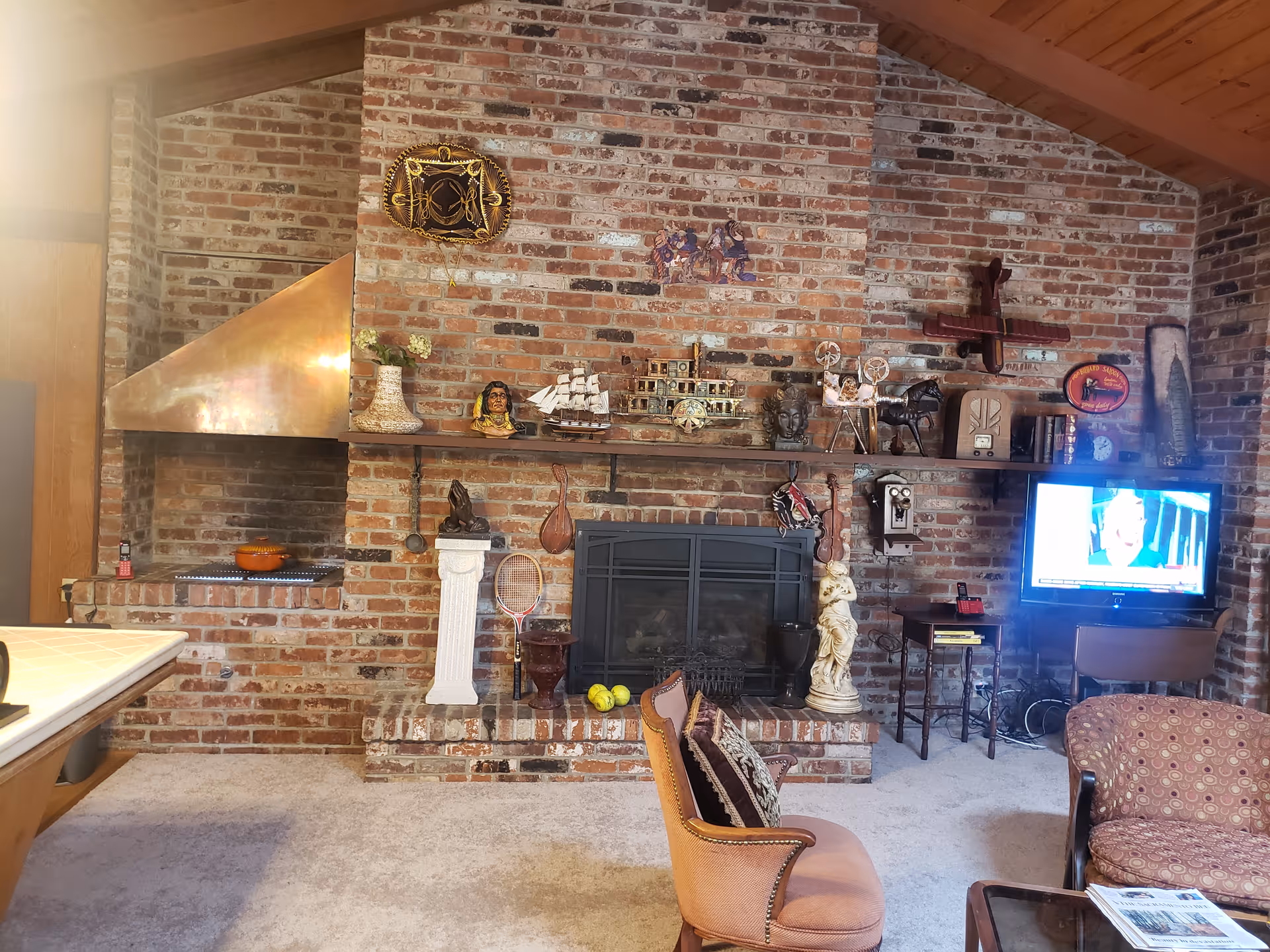 A cozy living room with a brick fireplace wall decorated with various ornaments including a ship model, busts, a tennis racket, and a statue. There is a TV on the right side showing a news program, a small table with a phone, and two upholstered chairs in the foreground. The ceiling is wooden and slanted.