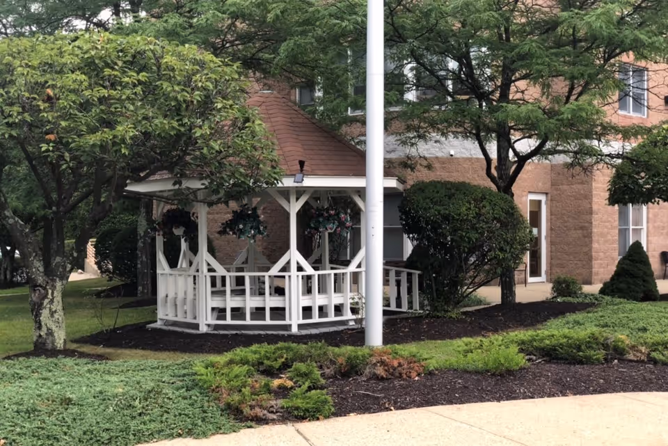 White wooden gazebo on landscaped grounds in front of a brick senior living facility building.