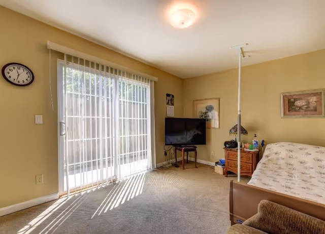 A cozy living room with beige walls and carpeted floor. There is a large sliding glass door with vertical blinds letting in natural light. A wall clock is mounted to the left of the door. In the corner, a flat-screen TV sits on a small table. Next to the TV is a wooden nightstand with a lamp, some bottles, and a radio. A bed with a floral bedspread is partially visible on the right side of the image. Two framed pictures hang on the walls.