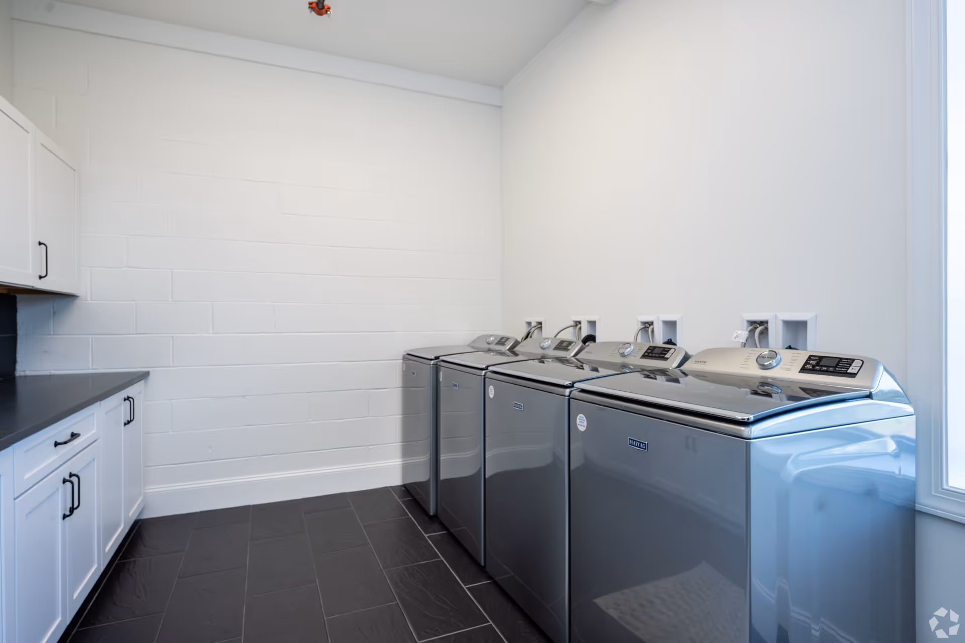 A clean laundry room with four modern silver washing machines lined up against a white wall. To the left, there are white cabinets with black handles and a black countertop. The floor is covered with dark tiles, and there is a large window on the right side letting in natural light.