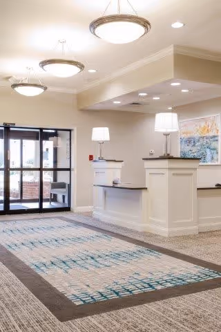 Bright and spacious reception area with a large patterned carpet, white reception desks with lamps, modern ceiling lights, and glass double doors leading outside.