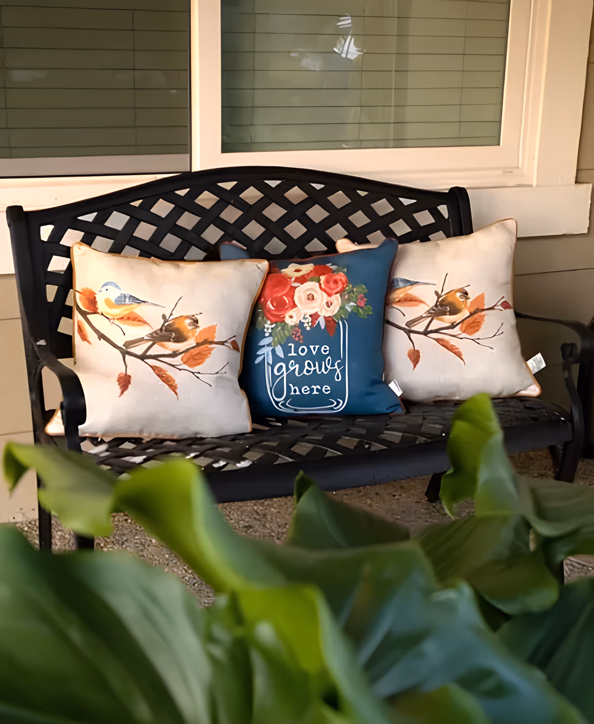 A black metal outdoor bench with a lattice backrest, adorned with three decorative pillows. Two pillows feature birds on branches with autumn leaves, and the middle pillow is blue with a floral design and the text 'love grows here'. The bench is placed in front of a window with closed blinds, and green leaves are visible in the foreground.