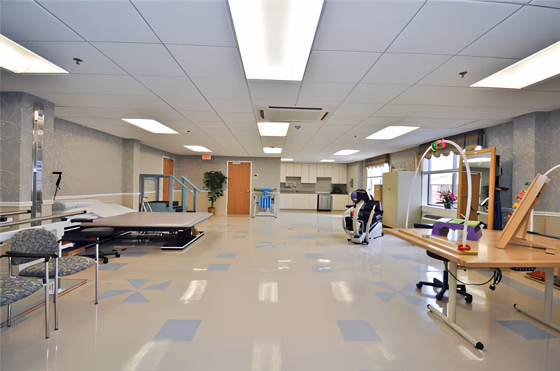 A spacious rehabilitation or therapy room with exercise equipment, therapy tables, chairs, and a kitchen area in the background. The room has a tiled floor with blue patterns, a drop ceiling with fluorescent lights, and large windows letting in natural light.