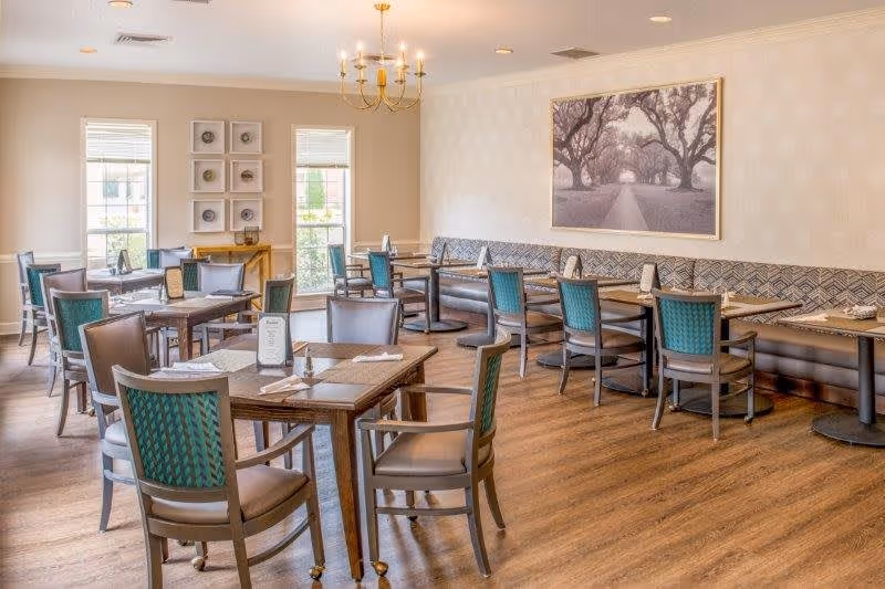 A bright and spacious dining room with multiple wooden tables and chairs arranged neatly. The room features large windows letting in natural light, a chandelier hanging from the ceiling, and a large framed black and white photograph of a tree-lined path on the wall. The floor is wooden, and there is a cushioned bench along one wall with chairs placed in front of it.