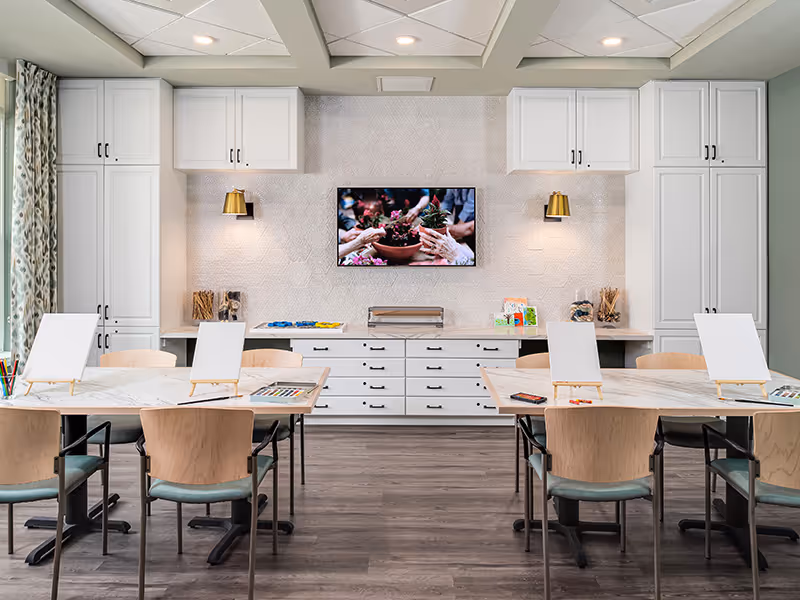 Bright activity room with tables, chairs and easels facing built-in white cabinetry and a wall-mounted TV.