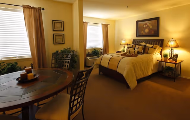 A warmly lit bedroom with a large bed dressed in beige and brown bedding, flanked by two bedside tables each with a lamp and small plants. There are two windows with beige curtains, a round wooden table with chairs, and framed artwork on the walls.
