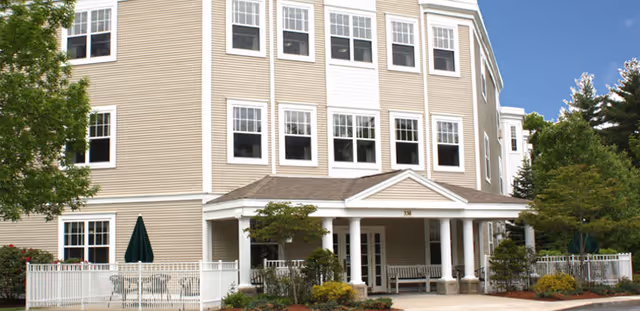 Exterior view of a multi-story senior living facility building with beige siding, white trim, multiple windows, a covered entrance with white columns, and landscaped greenery including trees and shrubs.