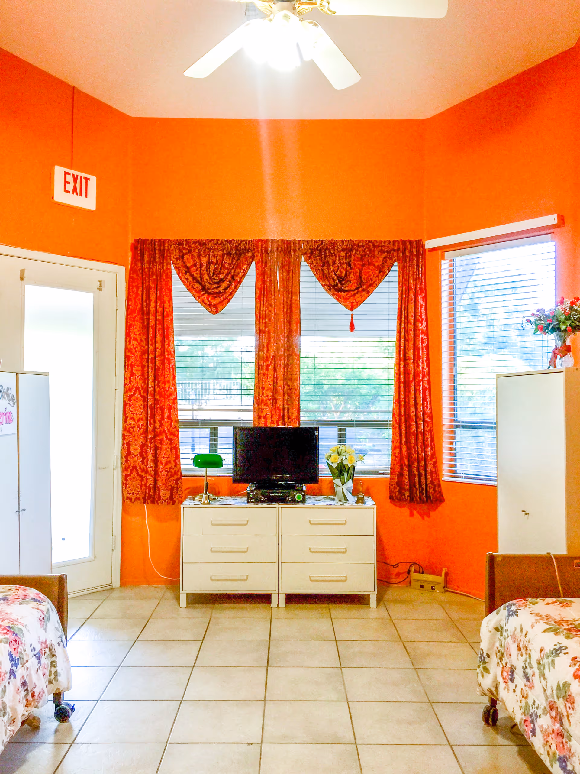 A bright room with orange walls featuring two single beds with floral bedspreads, a white dresser with a small TV, a green lamp, and a vase of flowers. There are large windows with red patterned curtains and blinds, a white ceiling fan, and a white door with an exit sign above it.