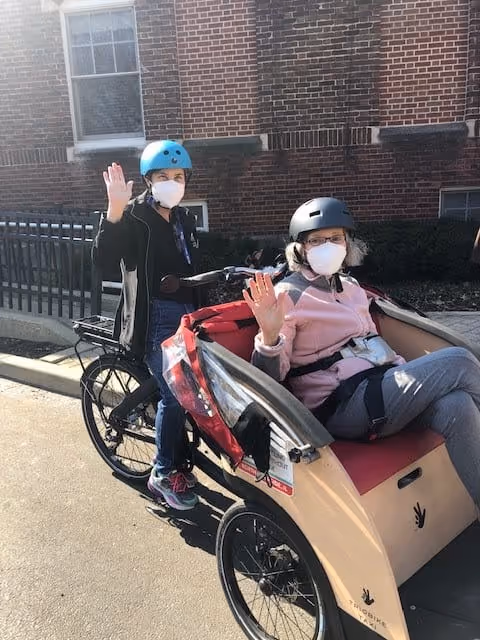 Two people outdoors near a brick building, both wearing helmets and face masks. One person is seated in a red and beige pedicab-style bicycle seat, while the other person stands behind holding the bicycle handles. Both individuals are waving at the camera.