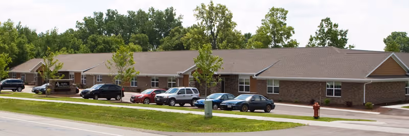 Exterior view of a single-story brick building with a brown roof, several windows, and a parking area in front with multiple cars parked. There are small trees and a grassy area along the sidewalk in front of the building under a partly cloudy sky.