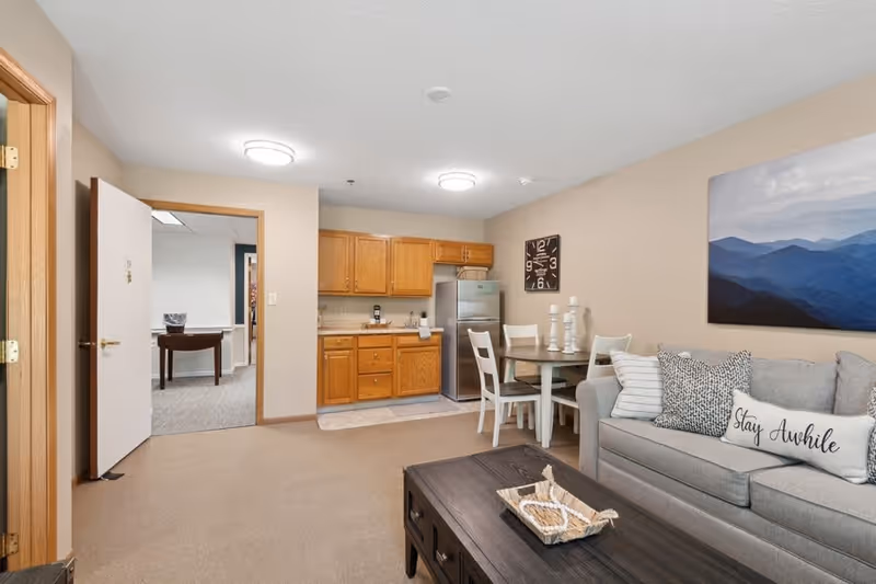 A cozy living area in a senior living facility featuring a gray sofa with decorative pillows, including one that says 'Stay Awhile'. There is a dark wooden coffee table with a small decorative basket on it. To the right of the sofa is a small round dining table with four white chairs and three white candle holders. The kitchen area has wooden cabinets, a stainless steel refrigerator, and a countertop with a coffee maker. A large wall clock and a landscape painting of blue mountains decorate the walls. An open door leads to another room with a table and chair visible.