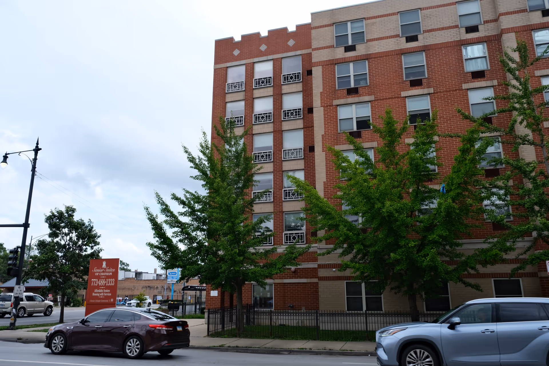 Front exterior of a multi-story red-brick senior living building with trees and cars along the street.