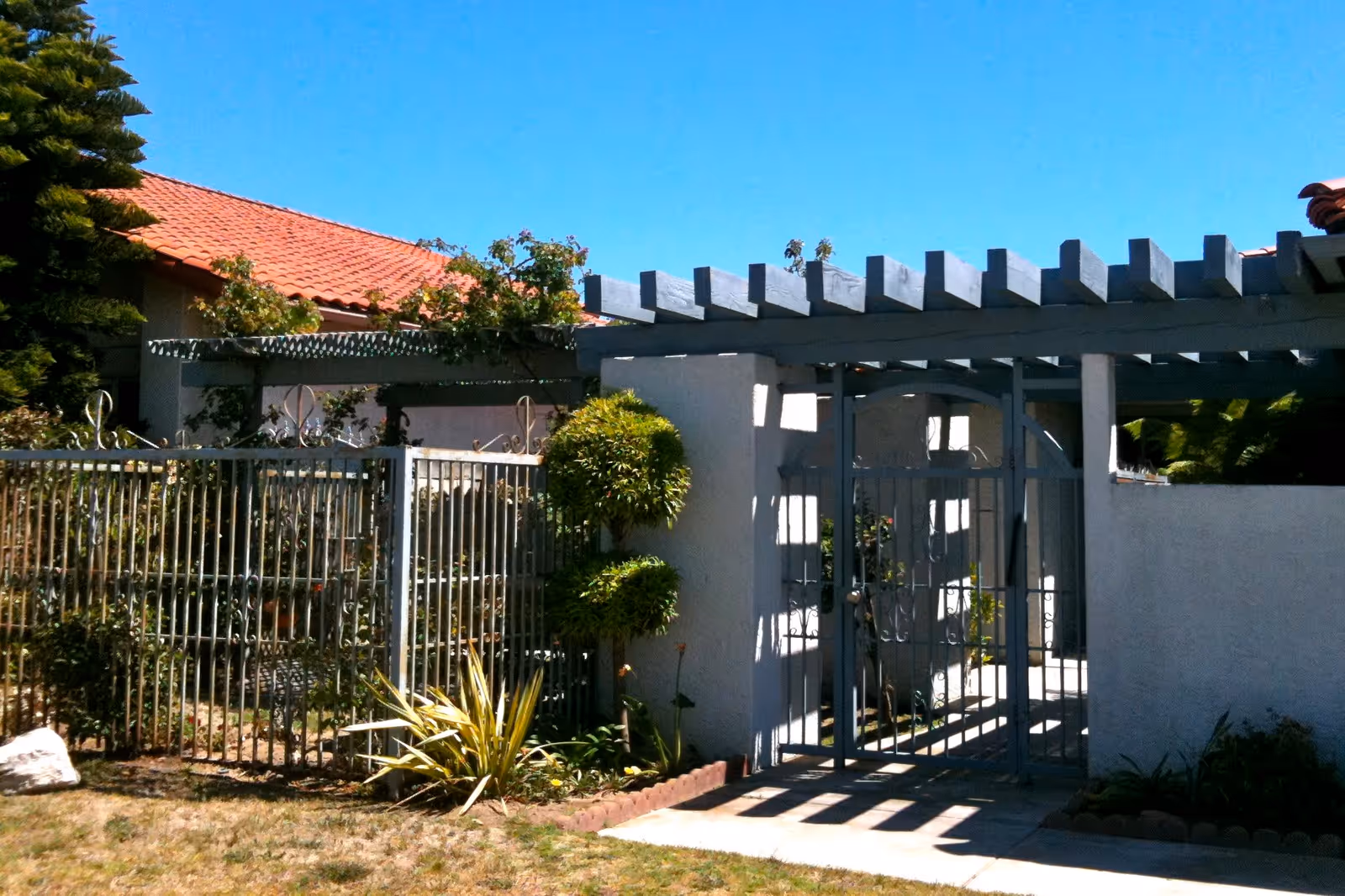 Outdoor view of a gated entrance with a metal fence and a pergola structure above the gate. There are trimmed bushes and plants around the entrance, and a building with a red tile roof is visible in the background under a clear blue sky.