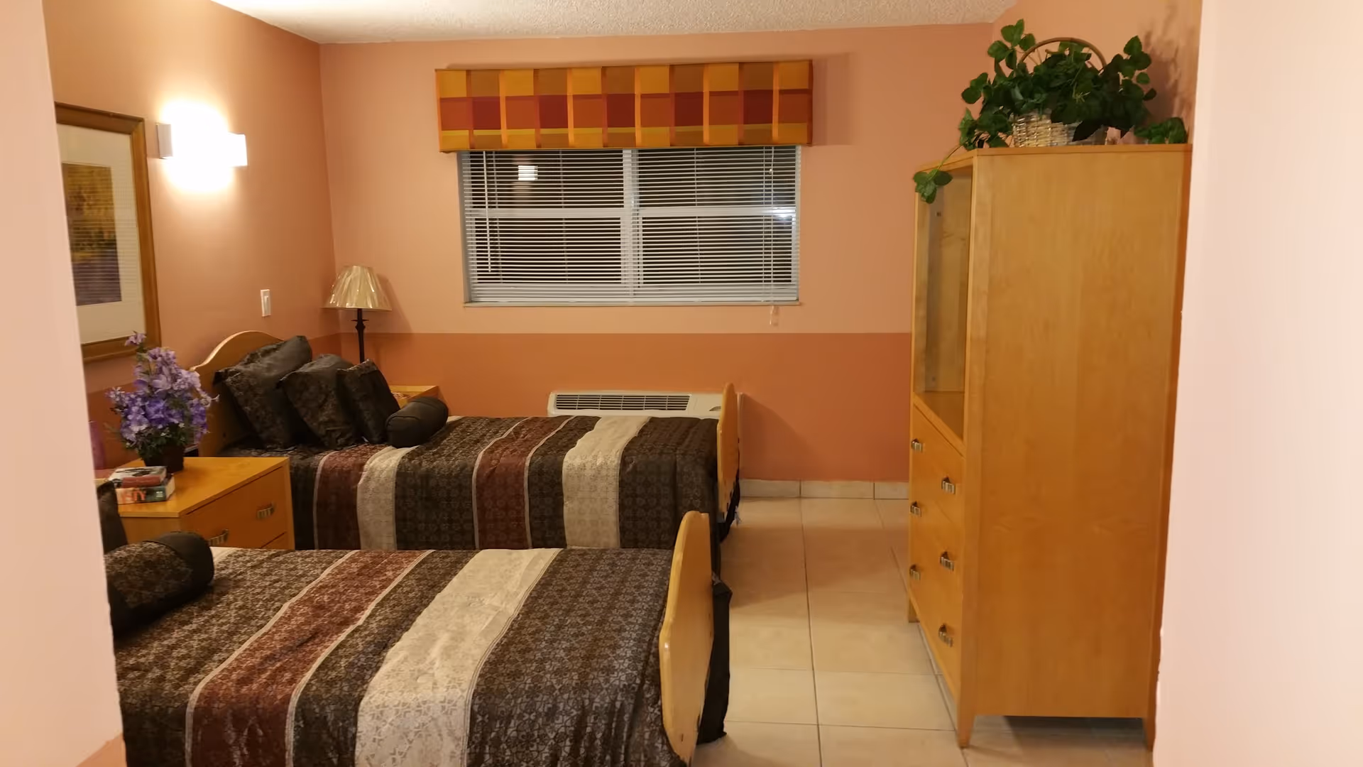 A bedroom with two single beds covered in patterned brown, beige, and maroon bedspreads. There is a wooden nightstand between the beds with a lamp and a vase of purple flowers. A framed picture hangs on the wall above the nightstand. A window with closed blinds and a colorful valance is centered on the far wall. On the right side of the room, there is a wooden dresser with drawers and a plant on top. The walls are painted in two shades of peach, and the floor is tiled.