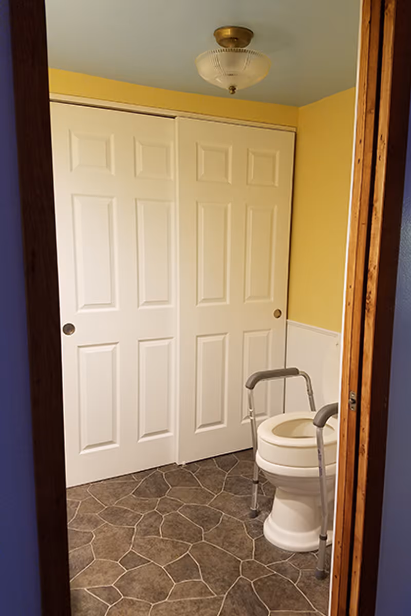 View into a bathroom with a toilet equipped with safety rails. The walls are painted yellow with white wainscoting, and there are white sliding closet doors on one side. The floor has a stone-patterned tile, and a ceiling light fixture is visible.