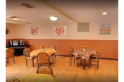 A communal dining room with several round tables and chairs, floral wall art, wood-paneled walls, and a small piano in the corner.