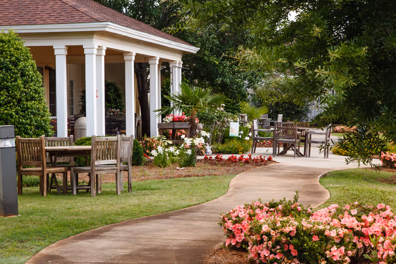 Curved concrete pathway winds through a landscaped garden with blooming flowers, wooden outdoor tables and chairs, and a covered porch with white columns.