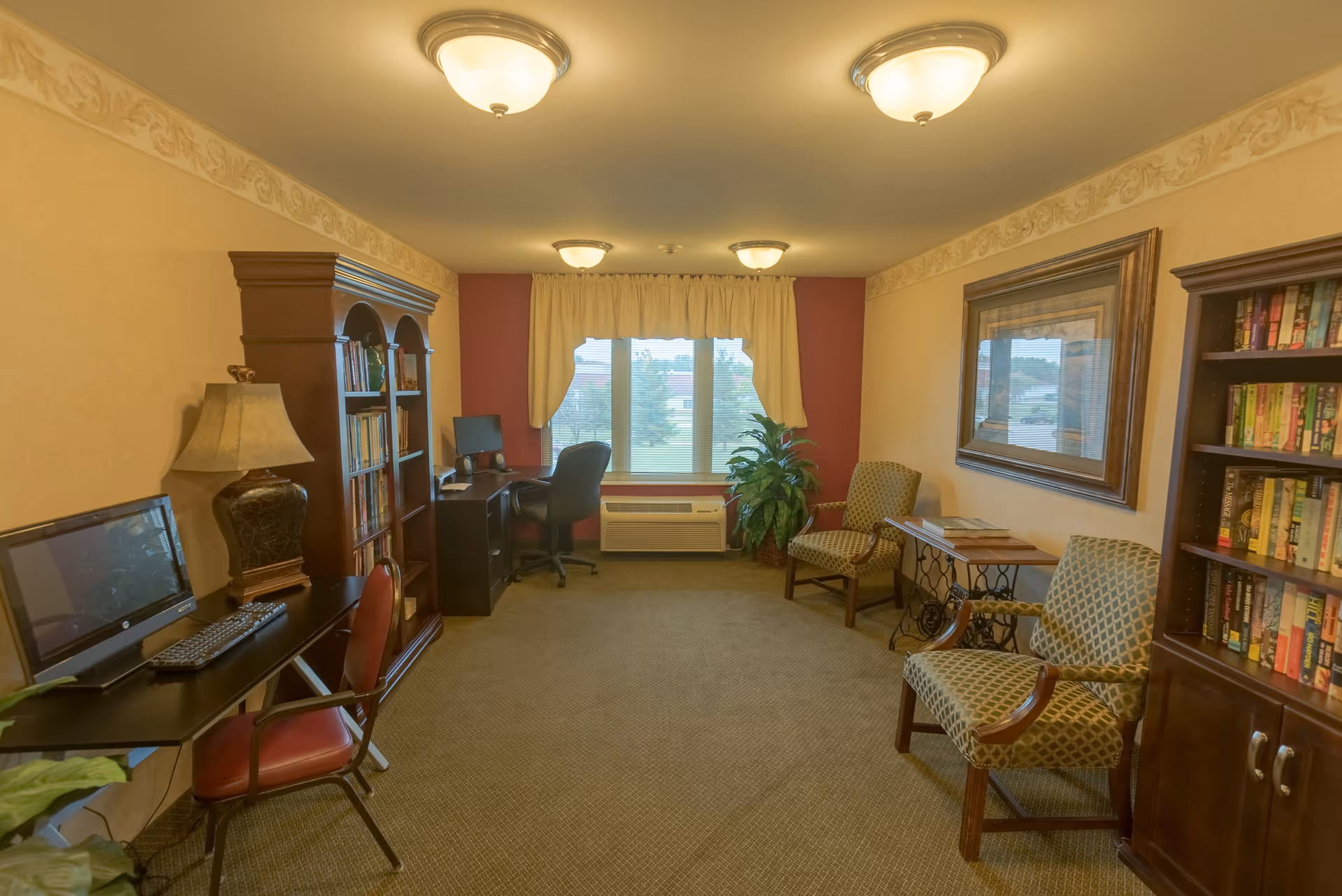 Well-lit common living room with bookshelves, computer desks, upholstered chairs and a window overlooking the outside.