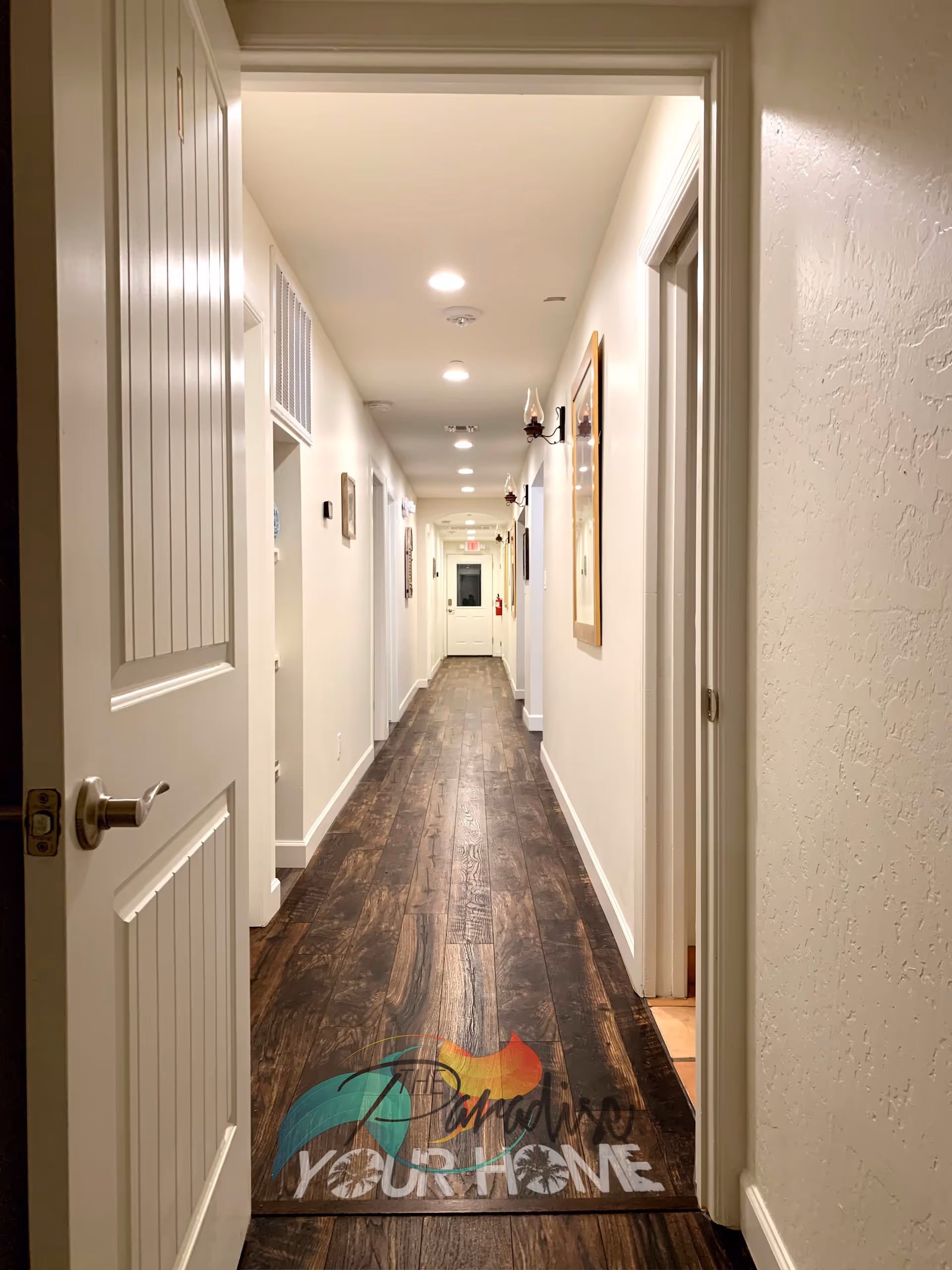 A long, well-lit hallway with wooden flooring and white walls. Several doors line both sides of the hallway, and framed pictures and wall-mounted lamps are visible on the right wall. The hallway leads to a white door at the far end with an exit sign above it.