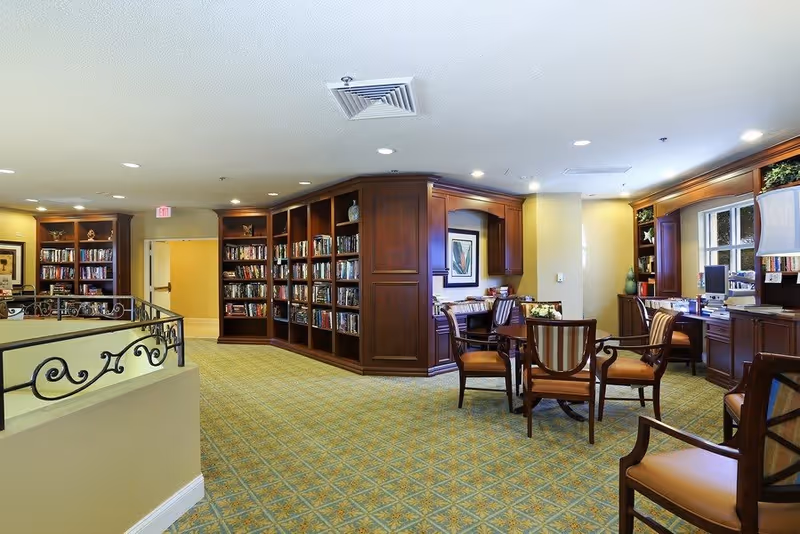 Interior view of a senior living facility library and common area with wooden bookshelves filled with books, a round table with four chairs, a desk with a computer, and warm lighting.