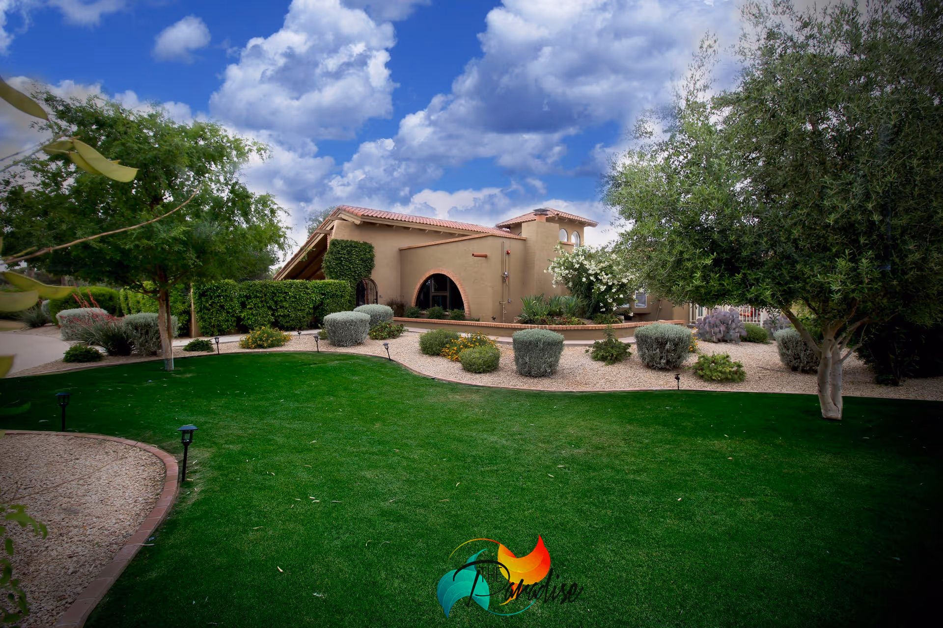 A well-maintained garden area with green grass, various shrubs, and trees in front of a beige stucco building with a red-tiled roof under a partly cloudy blue sky.
