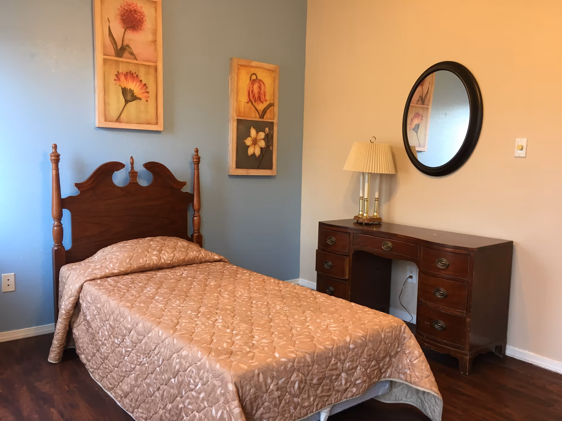 A bedroom with a single wooden bed covered with a beige quilted bedspread. On the wall behind the bed are two framed floral paintings. To the right of the bed is a wooden dresser with six drawers, a table lamp with a pleated shade, and a round mirror hanging on the wall above the dresser. The room has light blue and beige walls and a wooden floor.