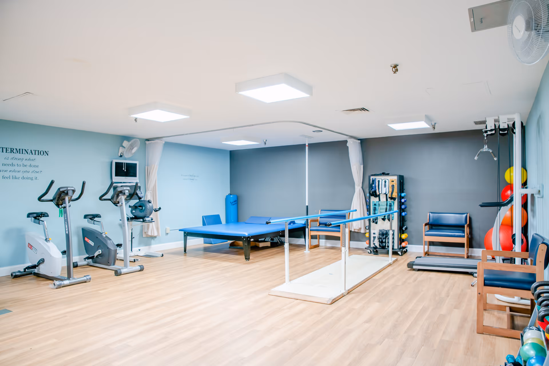 A rehabilitation or physical therapy room with exercise bikes, parallel bars for walking practice, a treatment table, chairs, and various exercise equipment including weights and exercise balls. The room has light blue and gray walls with wooden flooring and bright ceiling lights.