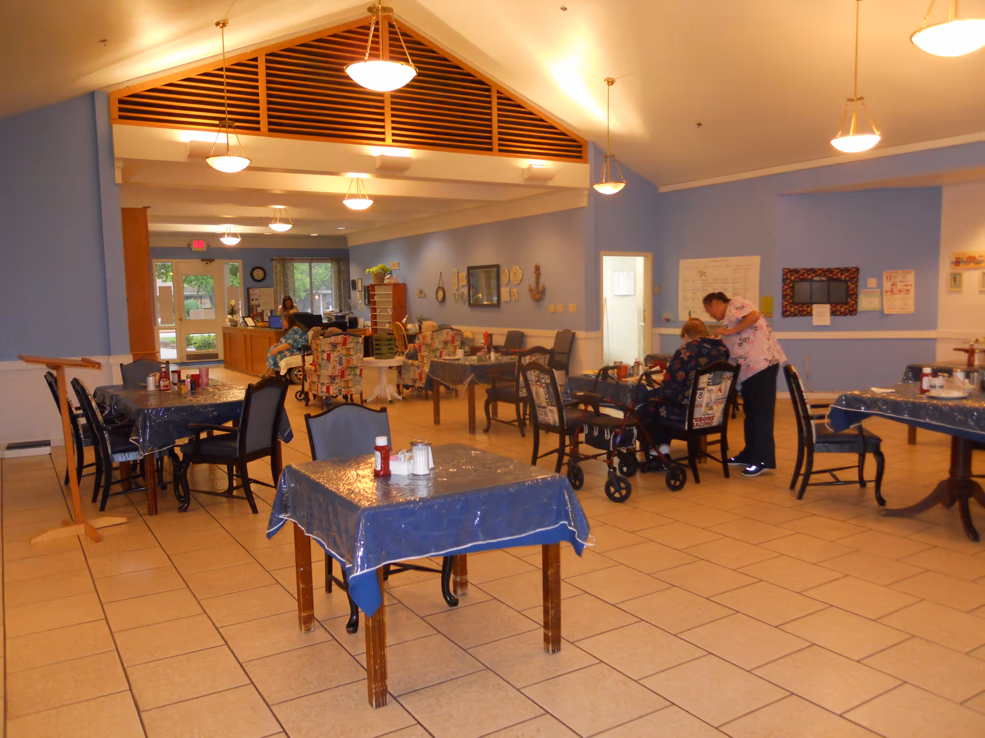 A spacious dining area in an assisted living facility with several tables covered in blue plastic tablecloths. There are chairs around the tables, and a few residents and a caregiver are visible. The walls are painted light blue, and there are ceiling lights hanging from a high ceiling. The room has tiled floors and a reception desk area in the background near the entrance.