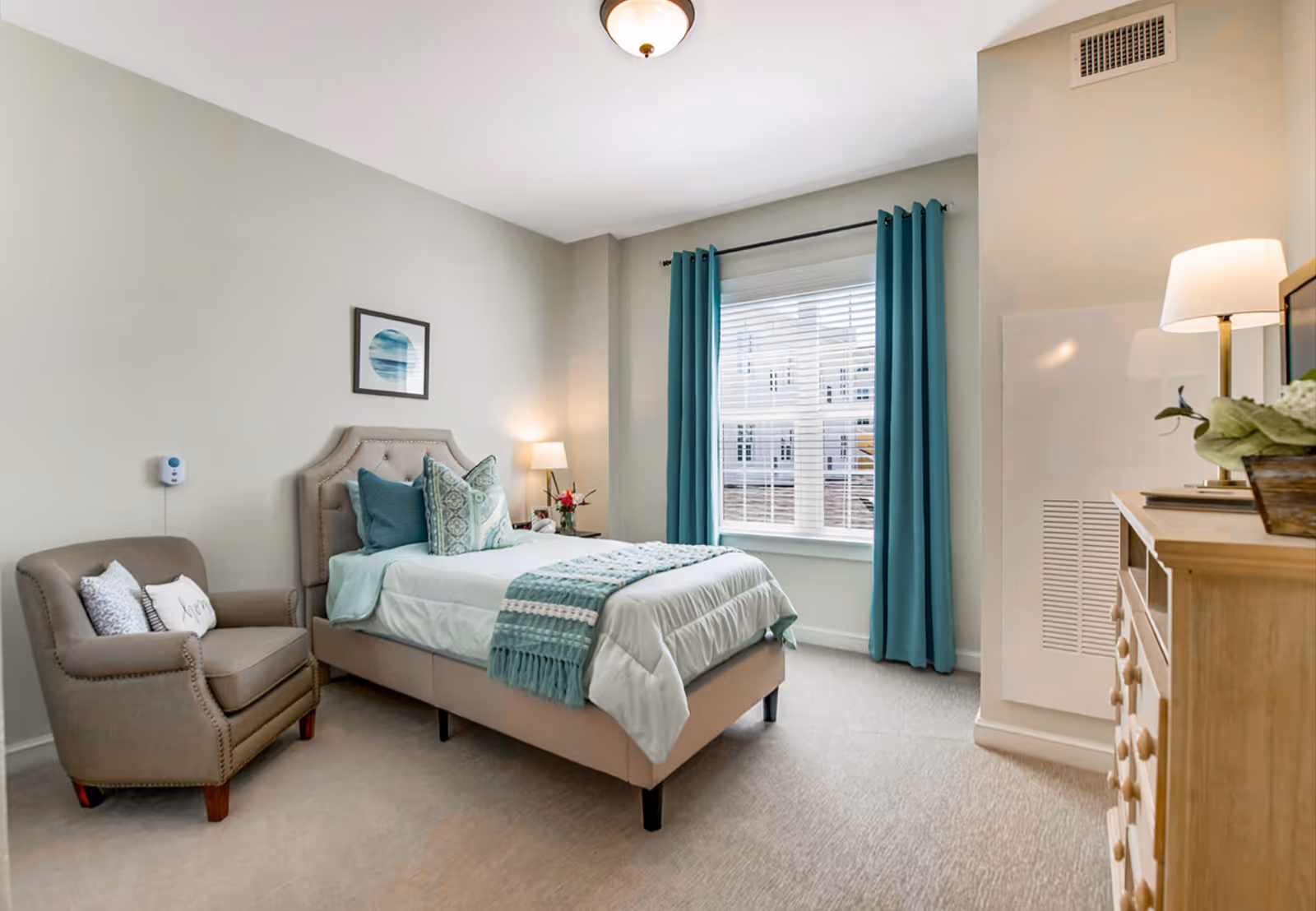 Bright furnished bedroom with a single bed, armchair, dresser, and teal curtains by a window.