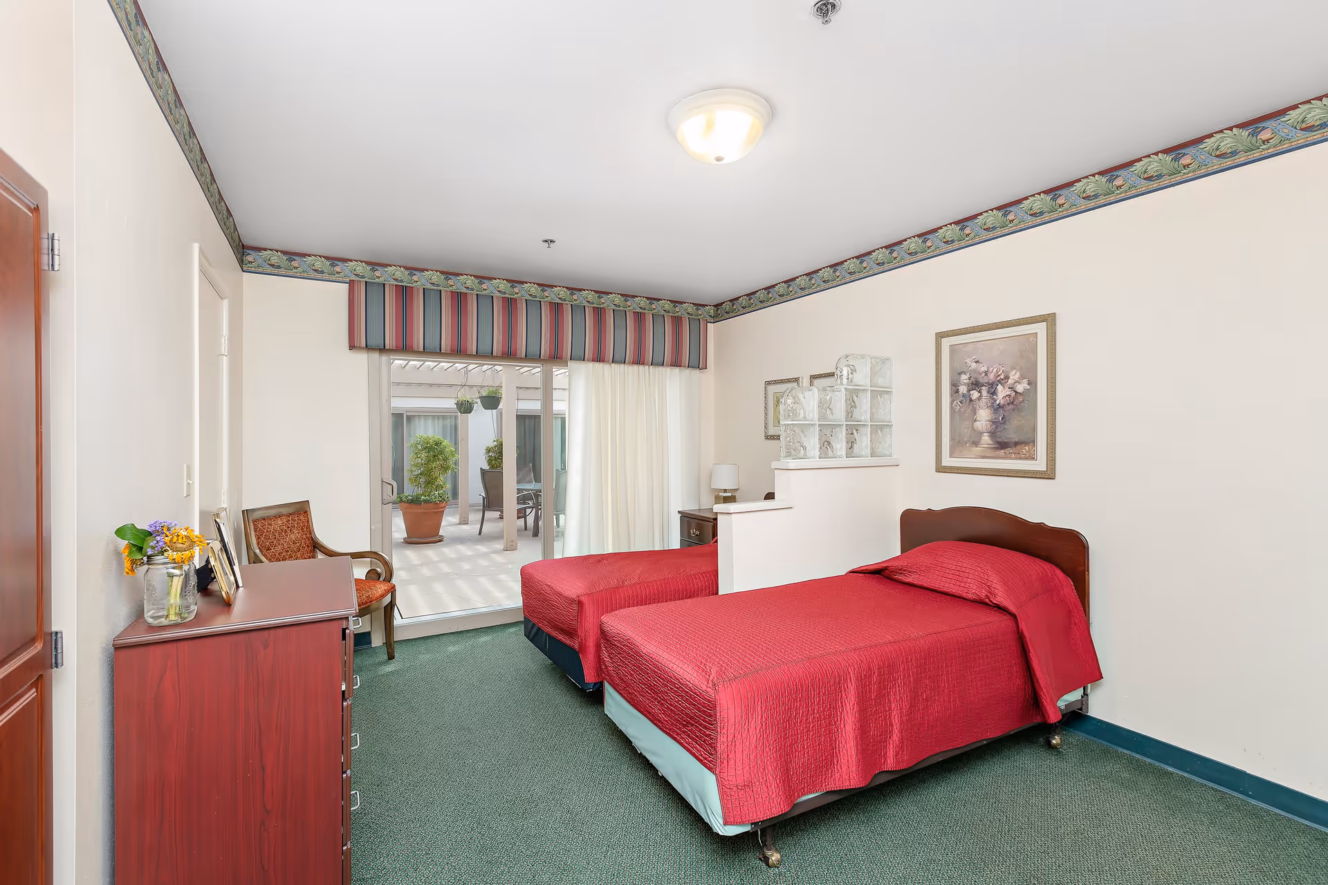 A bedroom with two single beds covered in red bedspreads separated by a half wall with glass blocks. The room has green carpet, a wooden dresser with a vase of flowers and picture frames, a chair, and a sliding glass door leading to an outdoor patio with potted plants. The walls are white with a decorative border near the ceiling and framed artwork.