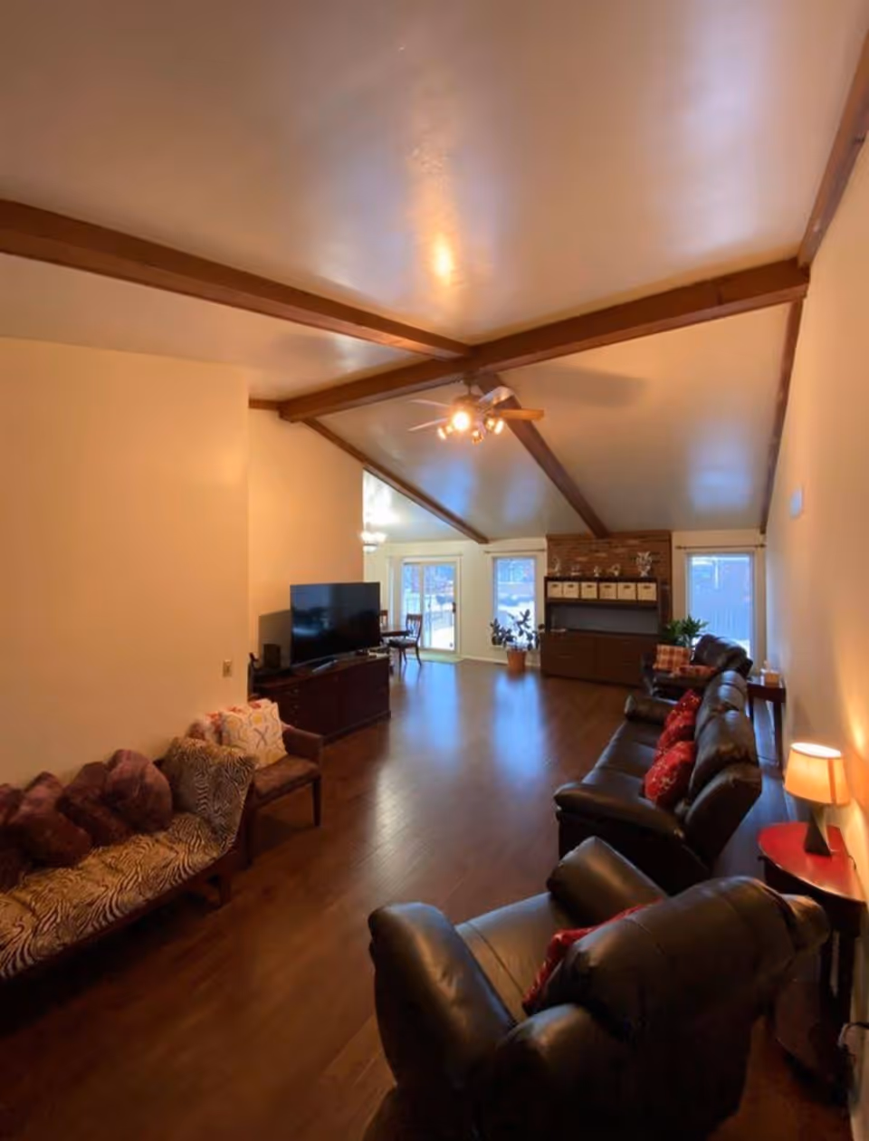 Spacious living room with a vaulted beamed ceiling, wood floors, a row of leather recliners, TV and windows at the far end.