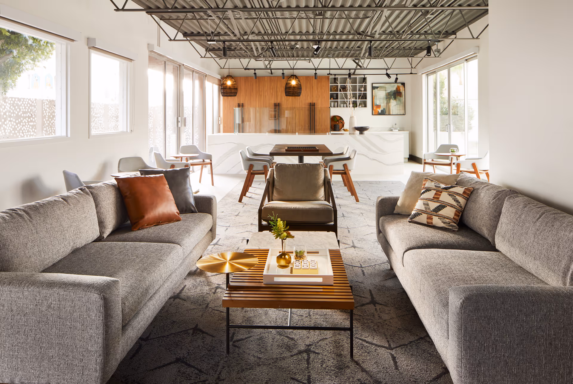 A modern living room with two gray sofas facing each other, a wooden coffee table with decorative items, a single armchair in the center, and a dining area with chairs and a table in the background. Large windows on both sides let in natural light, and the ceiling has exposed beams with track lighting.