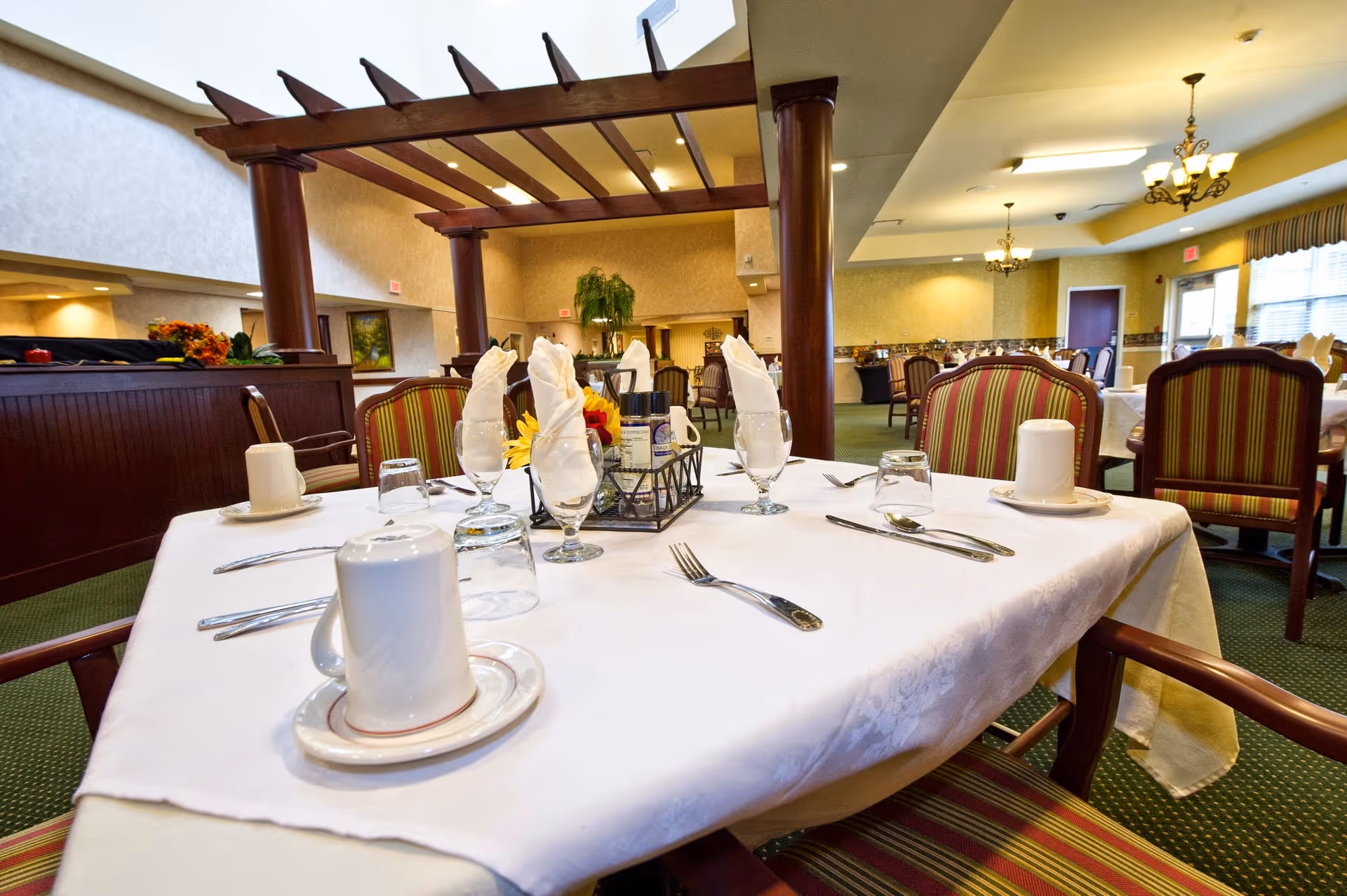 A dining room in a senior living facility with a table set for four. The table has a white tablecloth, folded white napkins in glasses, cups on saucers, silverware, and a centerpiece with flowers and condiments. The room features striped upholstered chairs, green carpet, chandeliers, and a wooden pergola structure in the background.