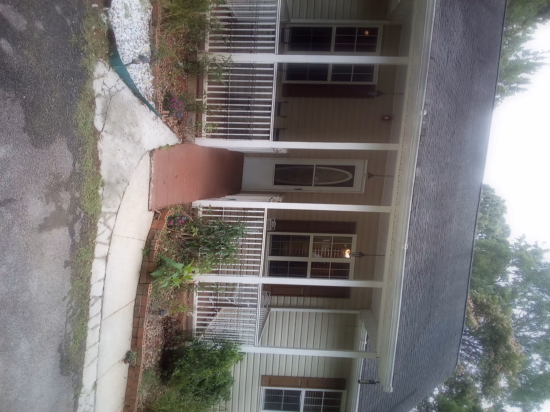 Front view of a single-story house with a small porch, white railing, and a ramp leading to the front door. The house has beige siding, brown shutters, and a shingled roof. There are plants and flowers along the walkway and porch area.