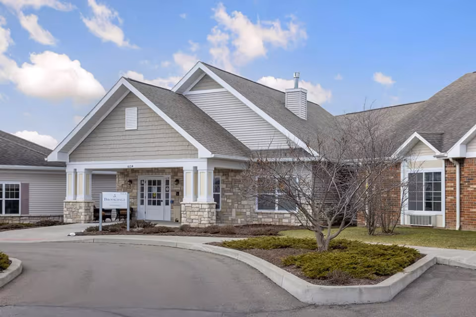 Exterior view of Brookdale Ithaca Assisted Living facility showing the entrance with a covered porch, stone and siding facade, a small landscaped area with bushes and a leafless tree, and a clear blue sky with some clouds.