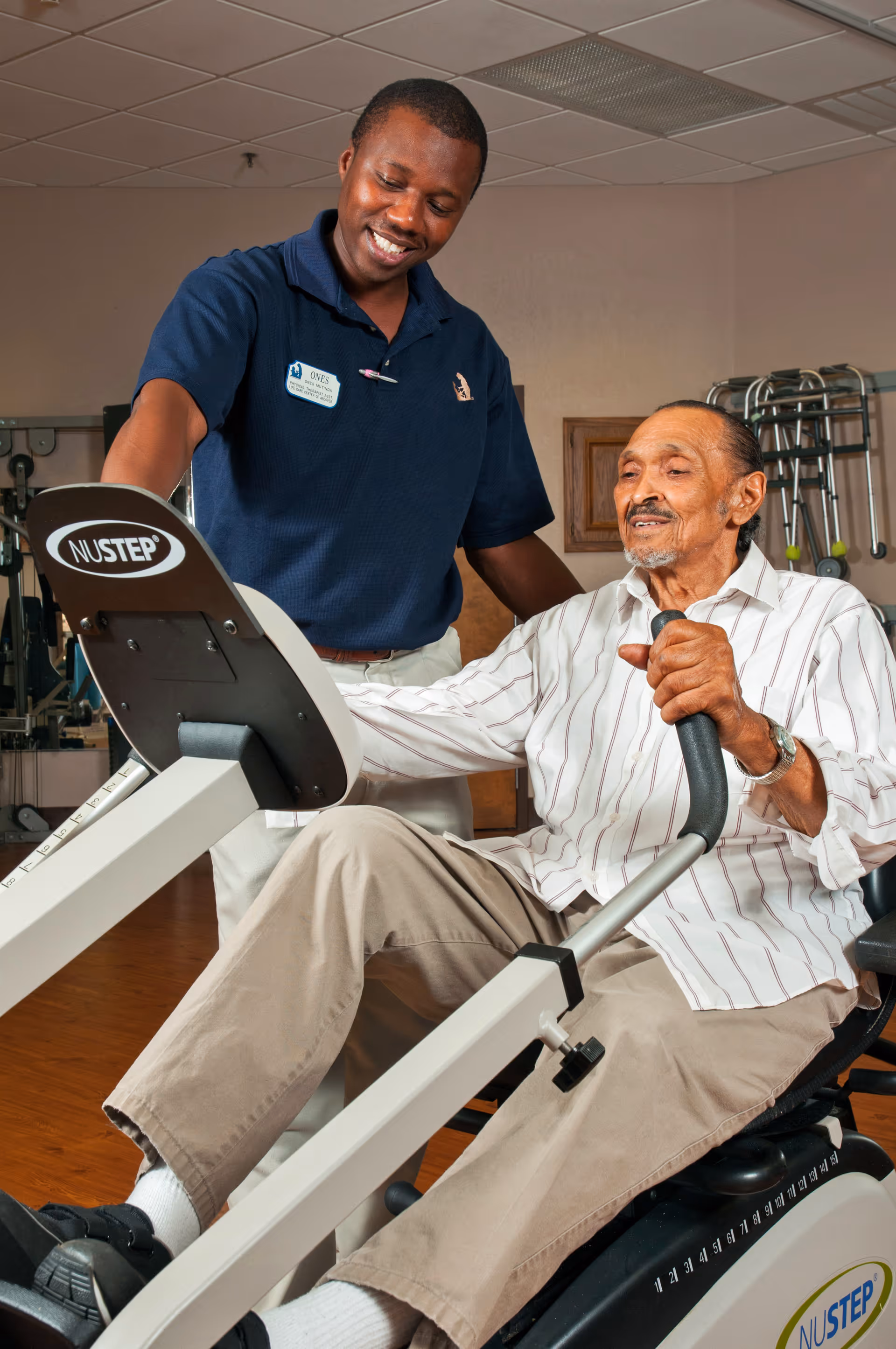 An elderly man using a NuStep exercise machine while a smiling caregiver or physical therapist stands beside him, offering support and encouragement in a fitness or rehabilitation room.