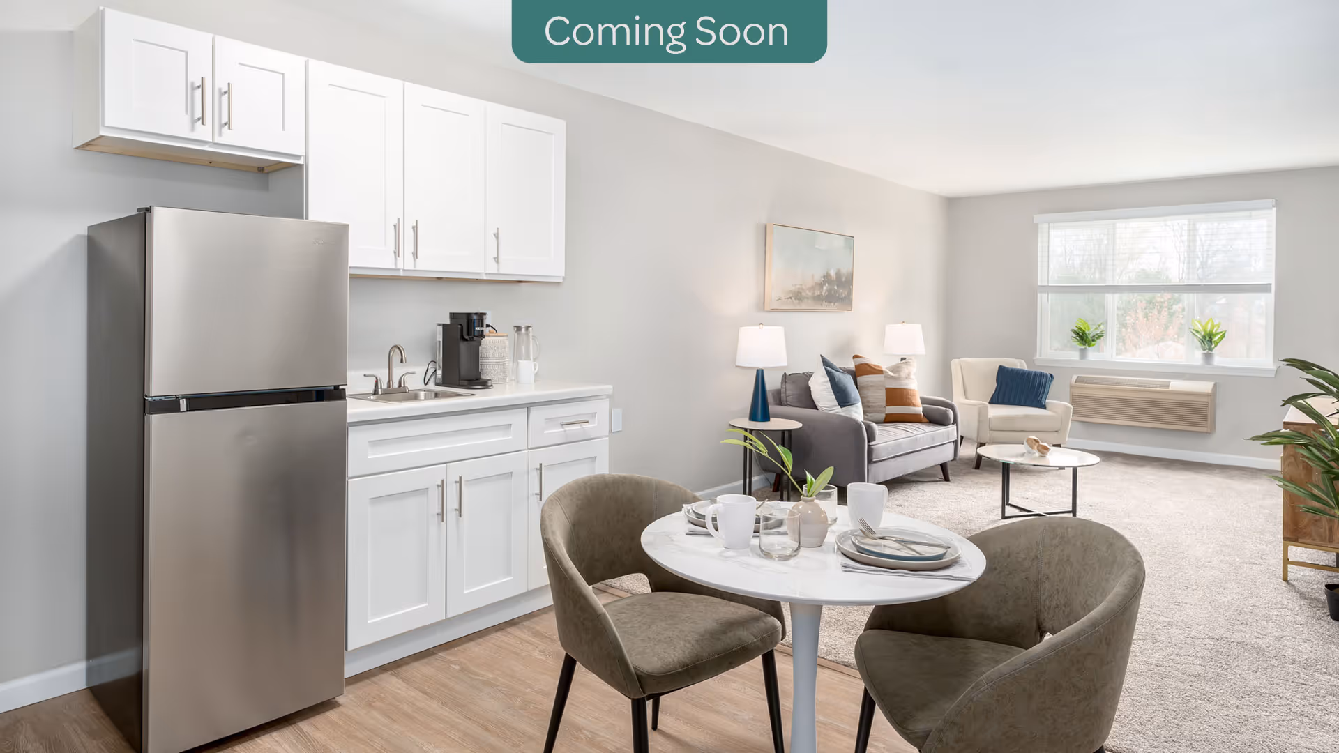 Bright open-plan living area with a small kitchenette, round dining table and chairs, sofa and armchair by a large window.