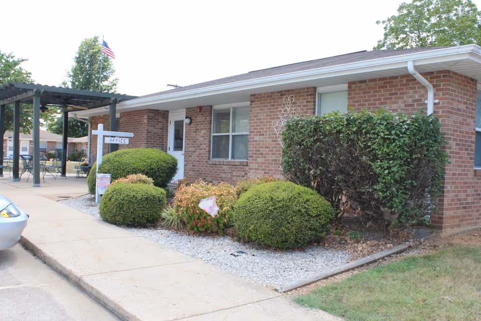 Exterior view of a single-story brick building with a white door and windows, surrounded by neatly trimmed bushes and a small garden bed with gravel. A white signpost near the entrance reads 'OFFICE'. There is a pergola with outdoor seating to the left and an American flag flying in the background.