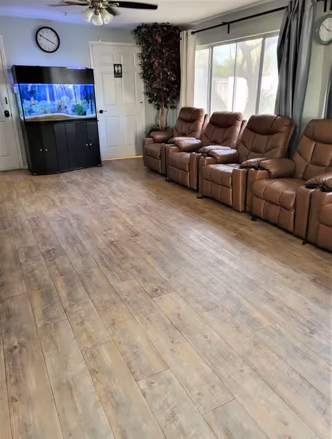 A bright room with four brown leather recliner chairs lined up against a wall with large windows covered by curtains. There is a large fish tank on a black stand near a white door with a restroom sign. A ceiling fan with lights is mounted on the ceiling, and a clock is visible on the wall above the fish tank. The floor is covered with light wood laminate.